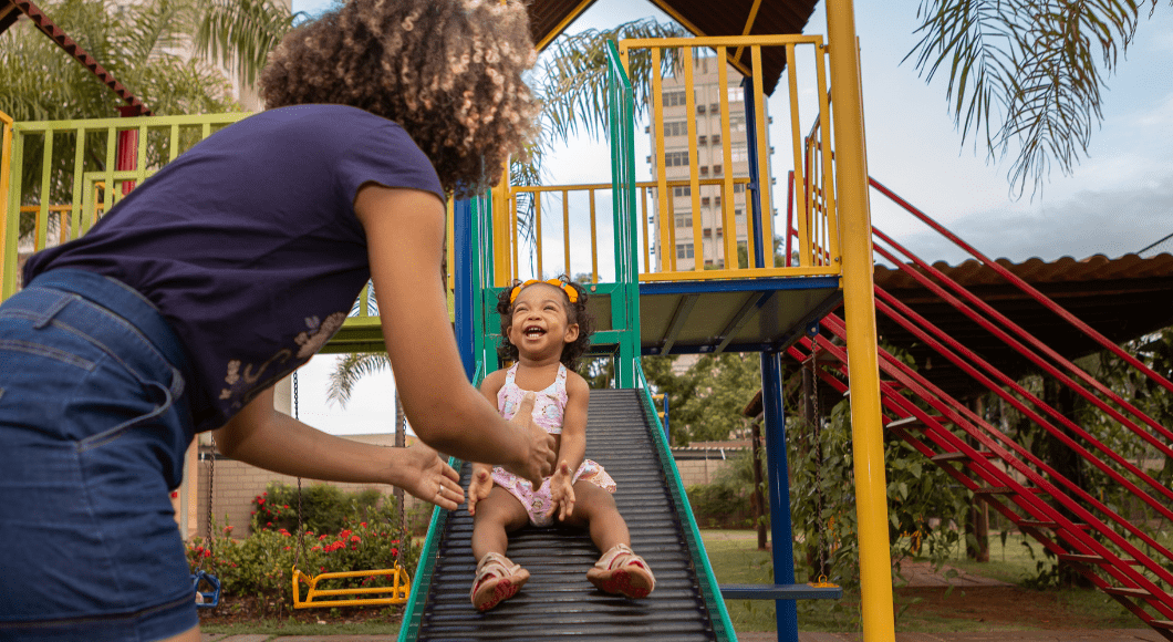 Toddler girl slides down roller slide into mama's arms at a park.