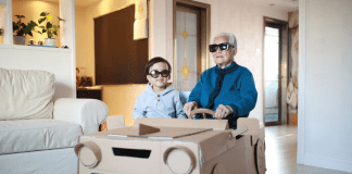 Grandpa and grandson sit in a homemade cardboard vehicle with sunglasses on.