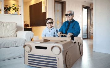 Grandpa and grandson sit in a homemade cardboard vehicle with sunglasses on.