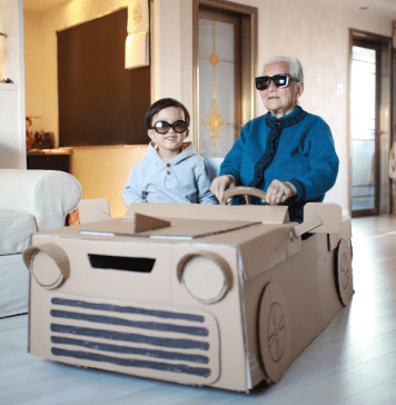 Grandpa and grandson sit in a homemade cardboard vehicle with sunglasses on.