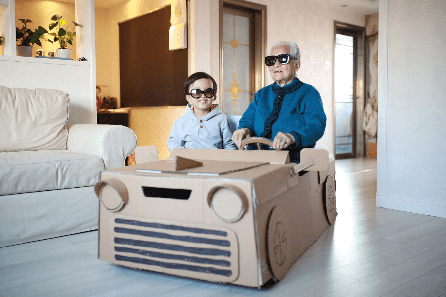 Grandpa and grandson sit in a homemade cardboard vehicle with sunglasses on.