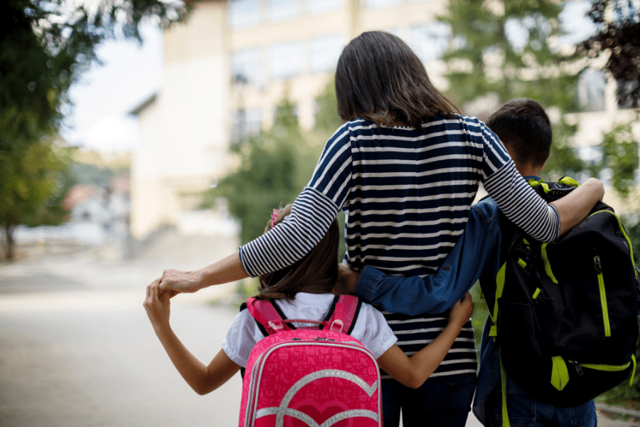 Black mom and two kids walk together to school down a sidewalk holding hands.