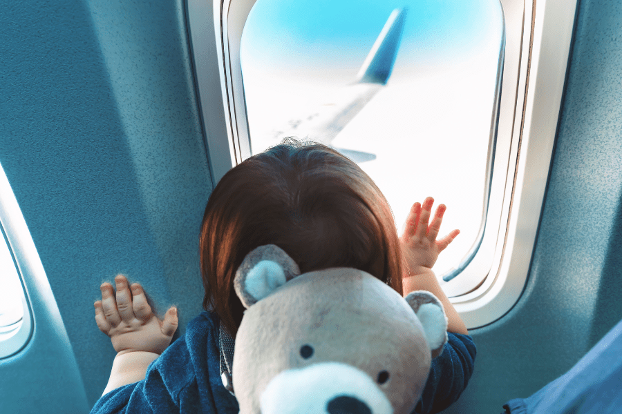 A toddler with a teddy bear backpack looks out the window of an airplane in flight.