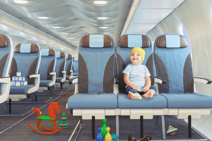 Toddler sits and smiles on an airplane with his toys strewn about the cabin floor.