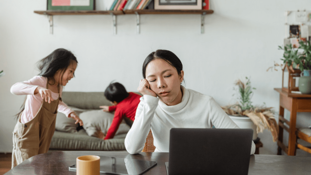 tired mom on laptop while children play behind her, exhaustion ITP