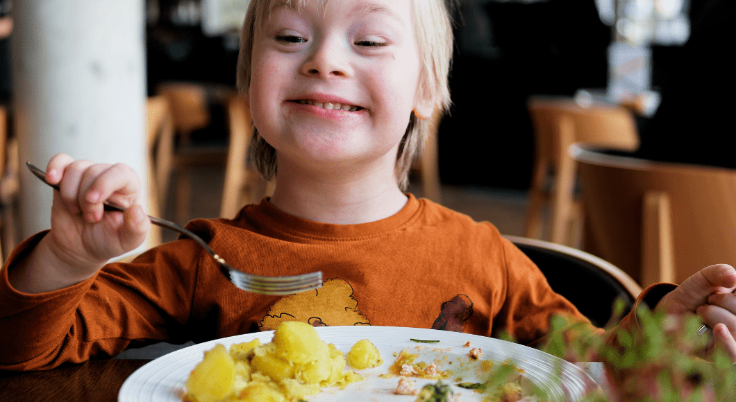 A little boy with Dow Syndrome in an orange shirt uses a fork to eat a meal while sitting in a chair at the table.