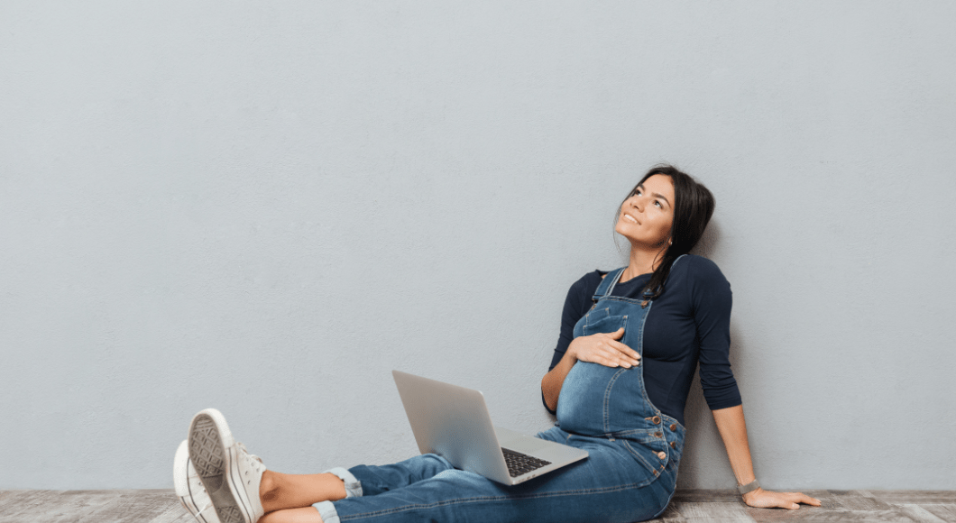 A pregnant woman sits on the ground with a laptop in her lap.