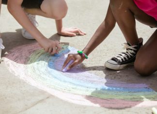 Two kids draw a rainbow with sidewalk chalk.