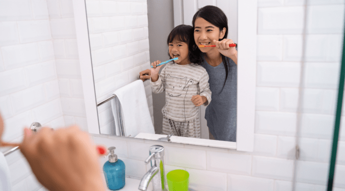 A mom and daughter brush their teeth in the bathroom mirror.