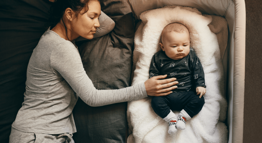 A tired mom lays next to and softly touches her baby in a bedside crib.