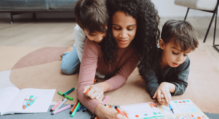 A mom and her kids draw in a coloring book together on the living room floor.
