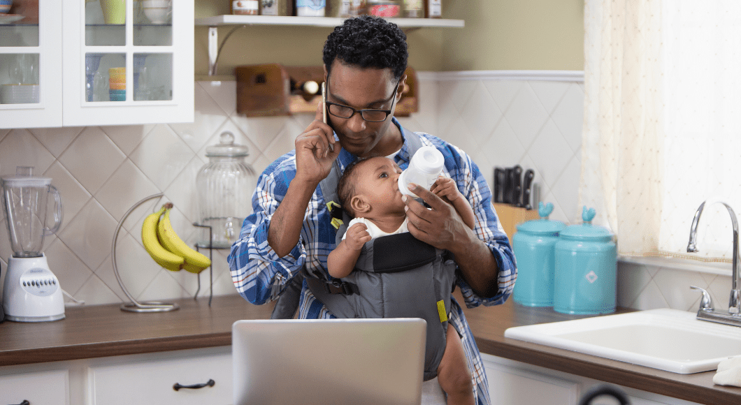 A father who is wearing baby in baby carrier and feeding bottle while looking at computer and on the phone