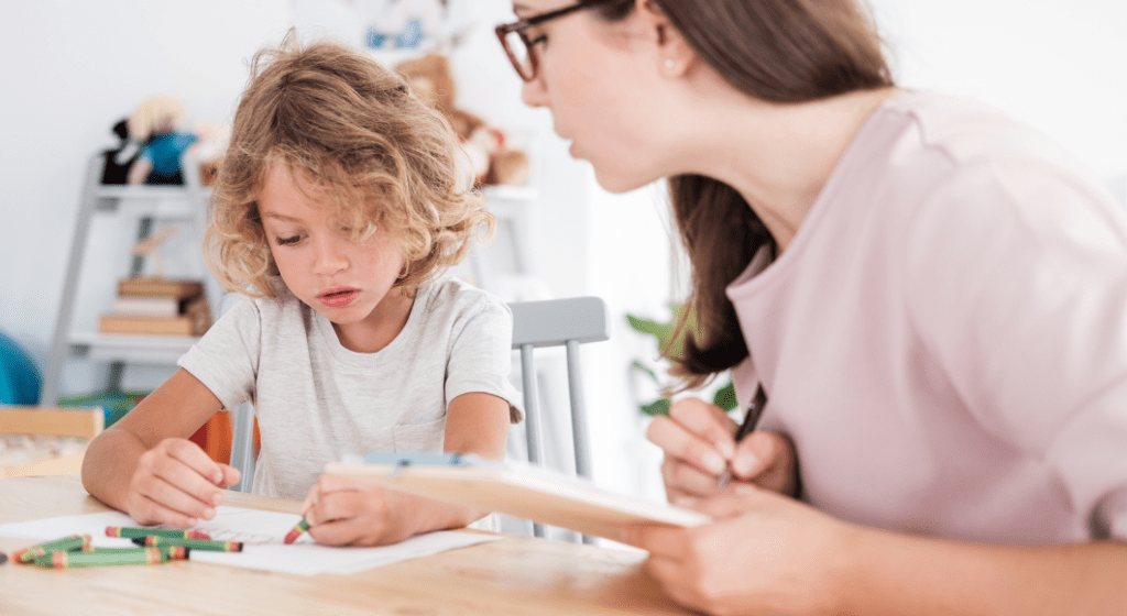 A woman who holding a clipboard sits next to a child coloring a picture.