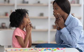 A speech pathologist models vowel sounds to a little girl who mimics her mouth shape.