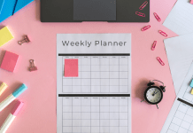 Weekly planner, markers, a clock, sticky notes, and a laptop sit on a pink pastel desk.