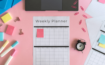 Weekly planner, markers, a clock, sticky notes, and a laptop sit on a pink pastel desk.