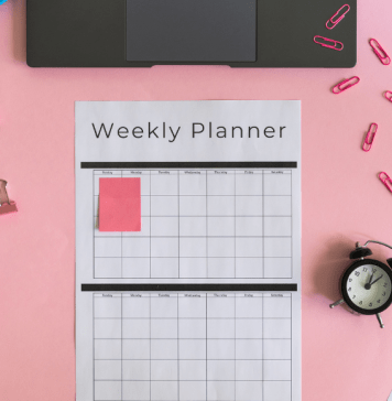 Weekly planner, markers, a clock, sticky notes, and a laptop sit on a pink pastel desk.