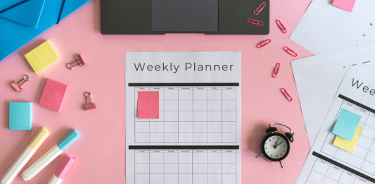 Weekly planner, markers, a clock, sticky notes, and a laptop sit on a pink pastel desk.