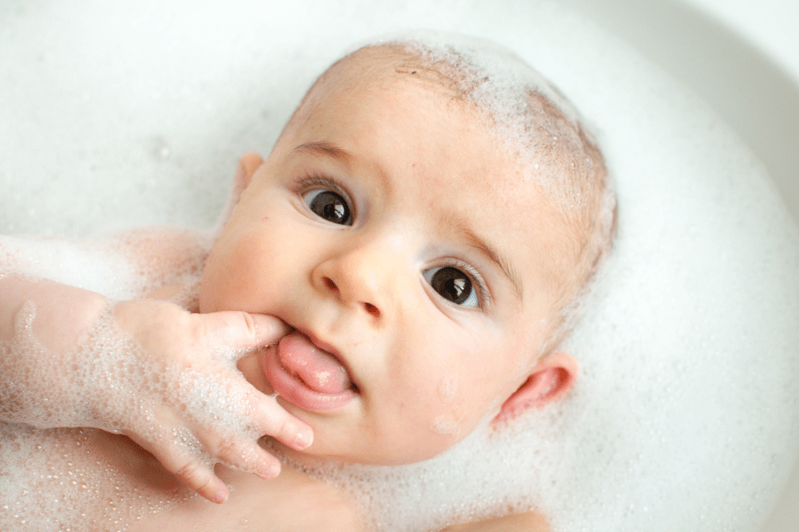 Baby lays in a soapy bath with a finger in his mouth, and looks surprised.