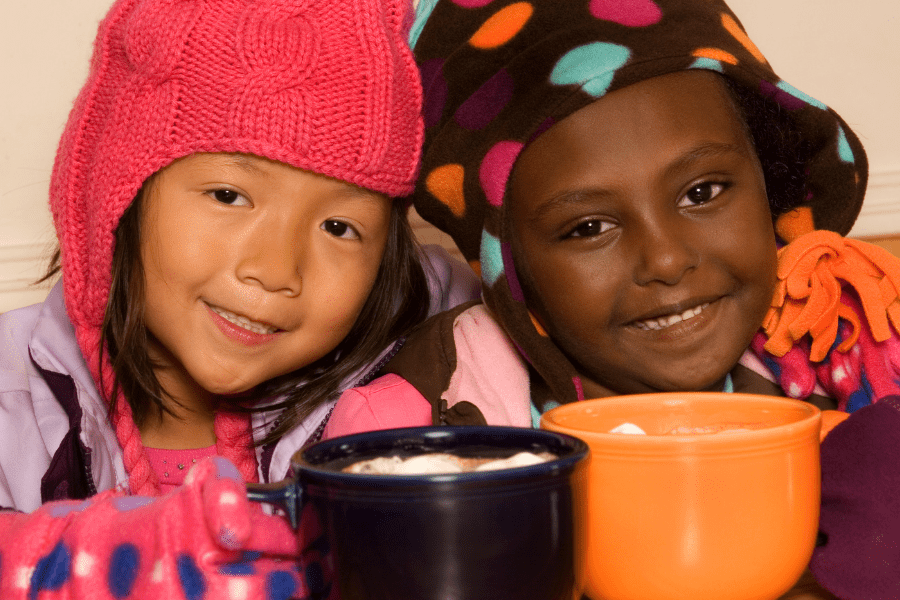 Two little girls wearing winter hats drink mugs of hot cocoa.