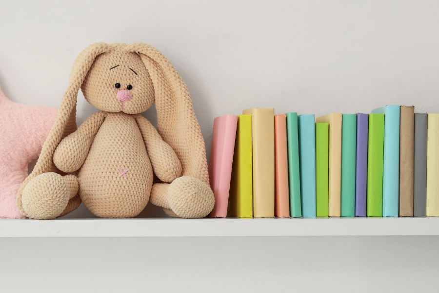 A bunny lovey with droopy ears sits on a bookshelf in a baby nursery.